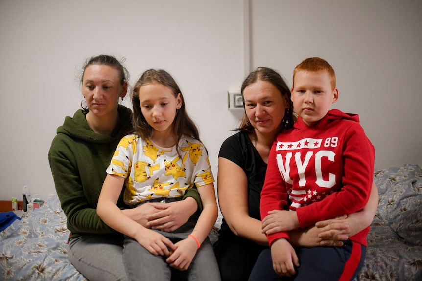 Two women sit on a bed. A young girl sits on one of the woman's laps. A boy sits on the other woman.