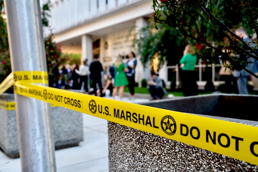 Yellow tape says US Marshal Do Not Cross with a group of people gathered behind it, in front of a court building.
