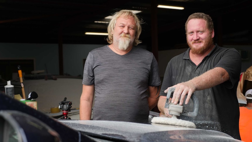 Two men stand together smiling while sanding an old car