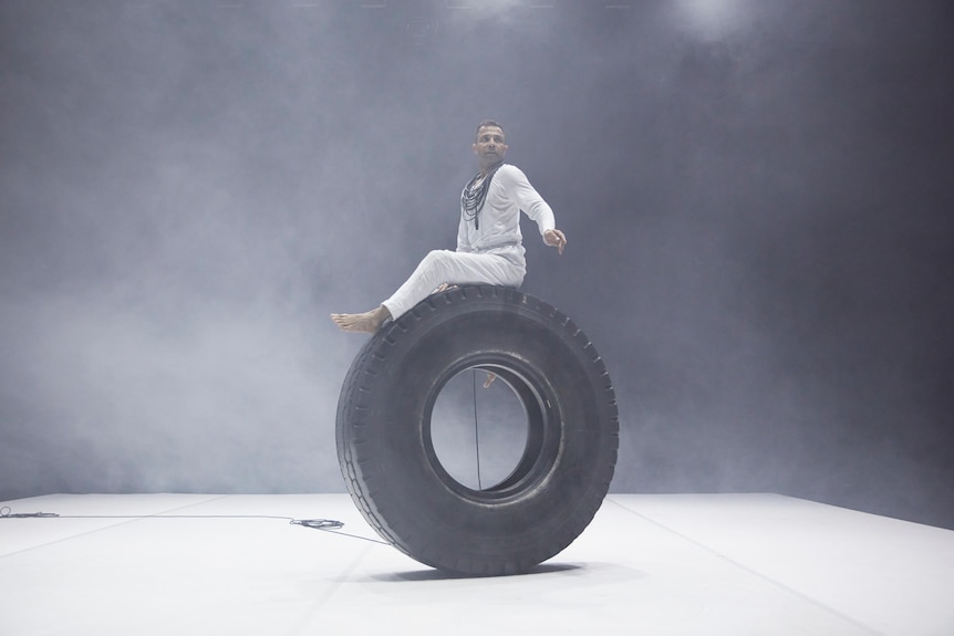 Indian-Australia man sits atop a large tyre on a stage wearing a shirt long-sleeved shirt and pants and surrounded by dry ice.