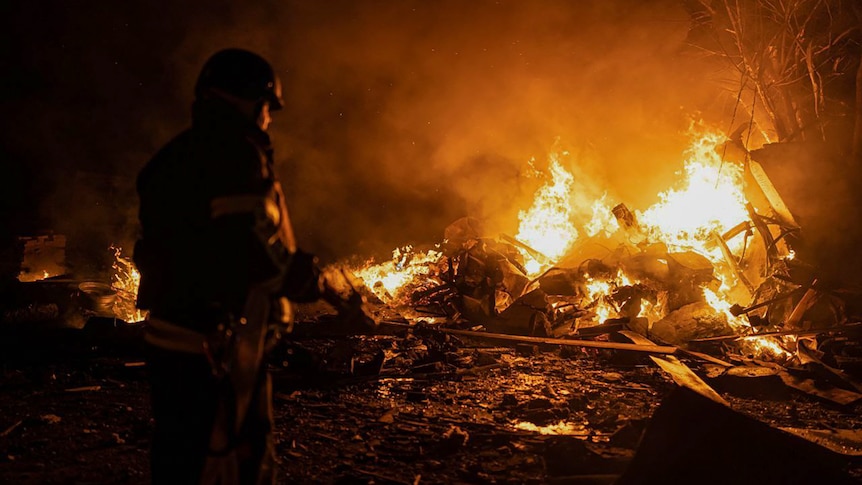 A person seen in silhouette wearing army gear as orange flames and fire burns from debris.