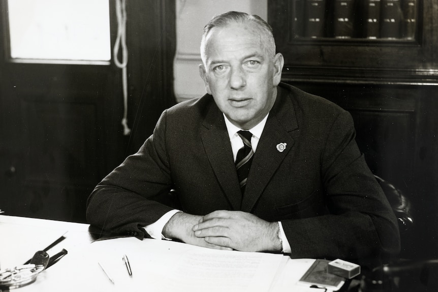 A man wearing a dark suit and tie sits at a desk with his hands folded.