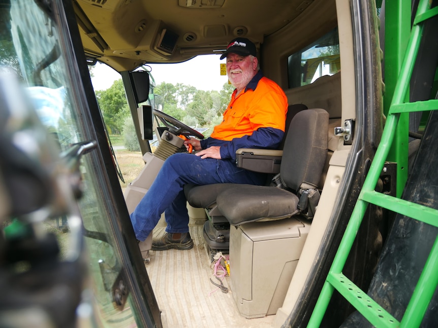 An older man with a beard, wearing a red and blue work shirt, smiles, while sitting in a cotton picker