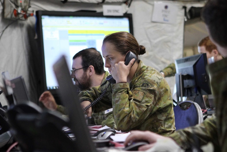 A woman in Australian Army uniform talks on the phone and looks at her computer in a room full of screens and computers.