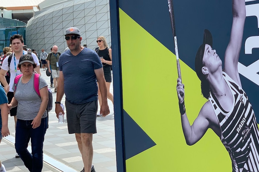 Tennis fans wearing caps, backpacks and sunglasses walk outside Rod Laver Arena on a sunny day past a poster of Ash Barty.