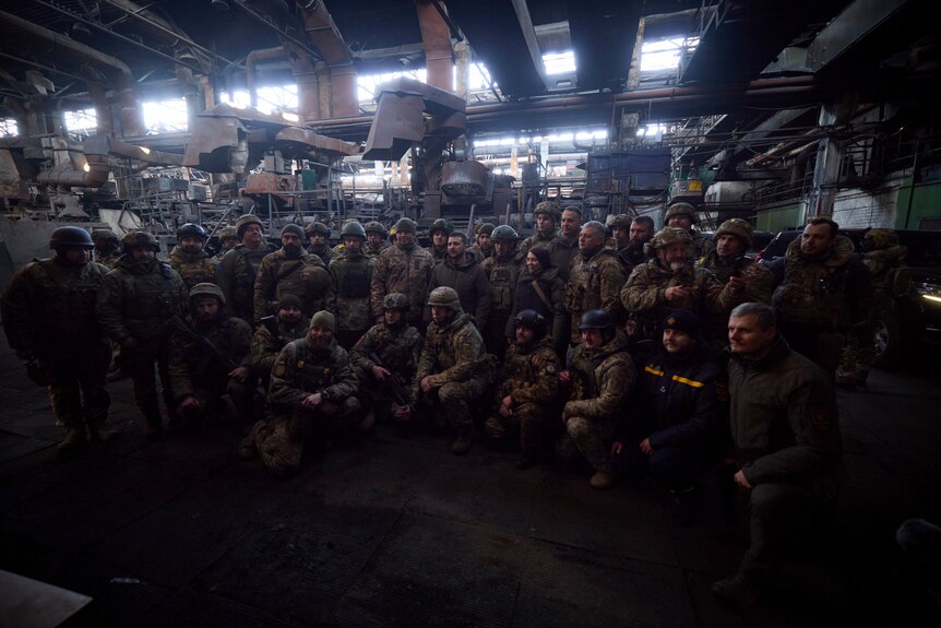A group of about 50 soldiers stand with a civilian politician in a dark building. 