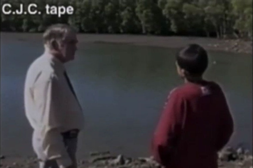 An older man listens to a young boy speaking, as both stand near a creek with bushland opposite.