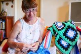 A woman sitting on a couch surrounded by crochet blankets, smiling and the camera looking over her glasses