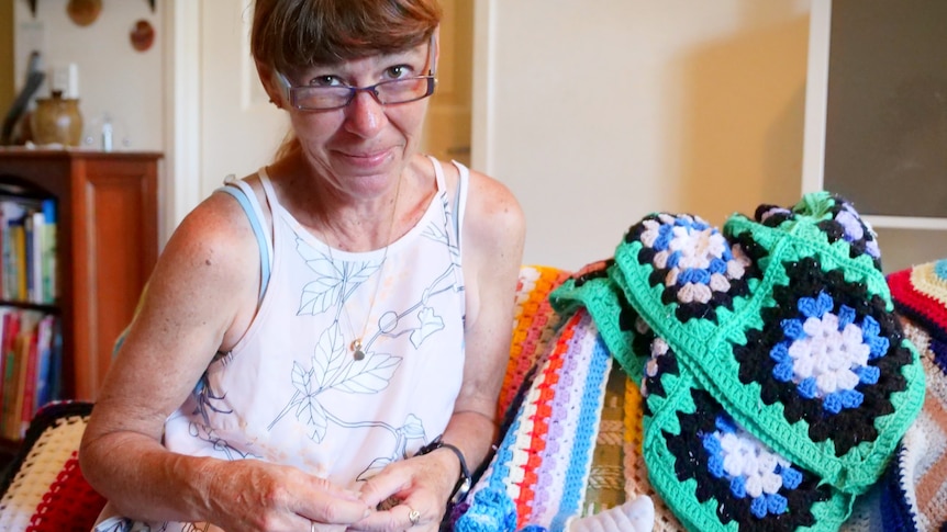 A woman sitting on a couch surrounded by crochet blankets, smiling and the camera looking over her glasses