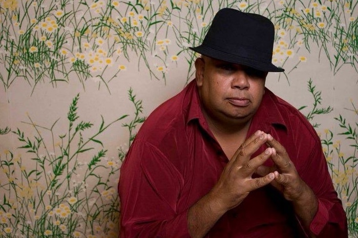 A man wearing a black hat and a red shirt sits in front of a wall painted with flowers.