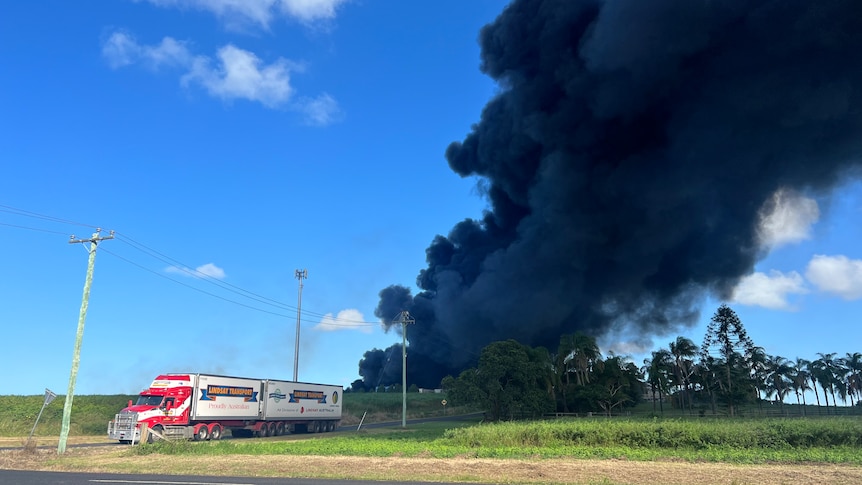 A truck drives along a road away from billowing black smoke in the distance.