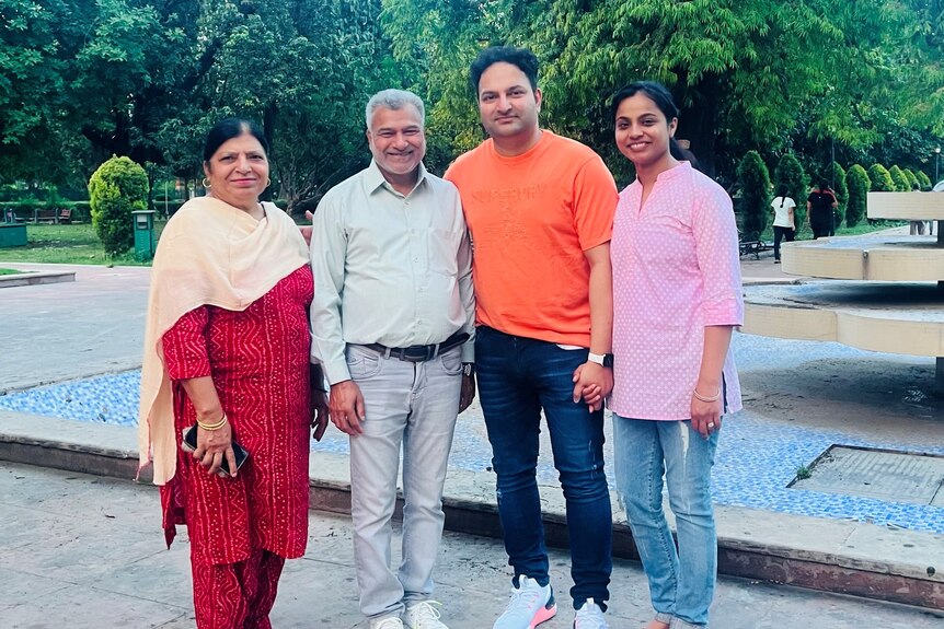 Four Indian people — a middle-aged couple and a younger couple — stand in front of a concrete fountain and trees