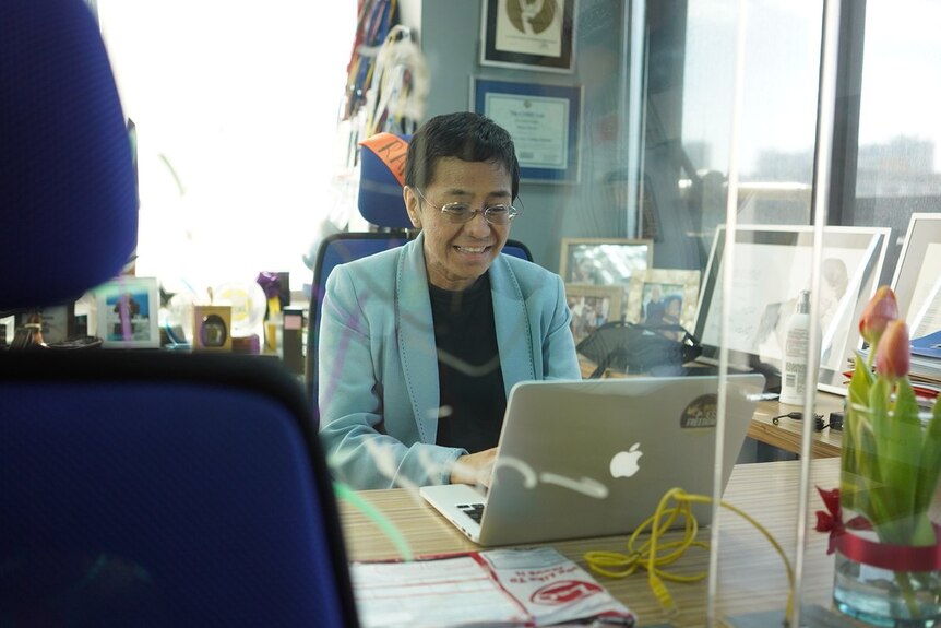 A woman in a suit jacket sits at a desk in a office and works on a laptop while smiling.