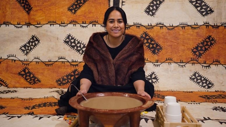 Tongan woman sits infront of a kava bowl smiling on a fine mat. 