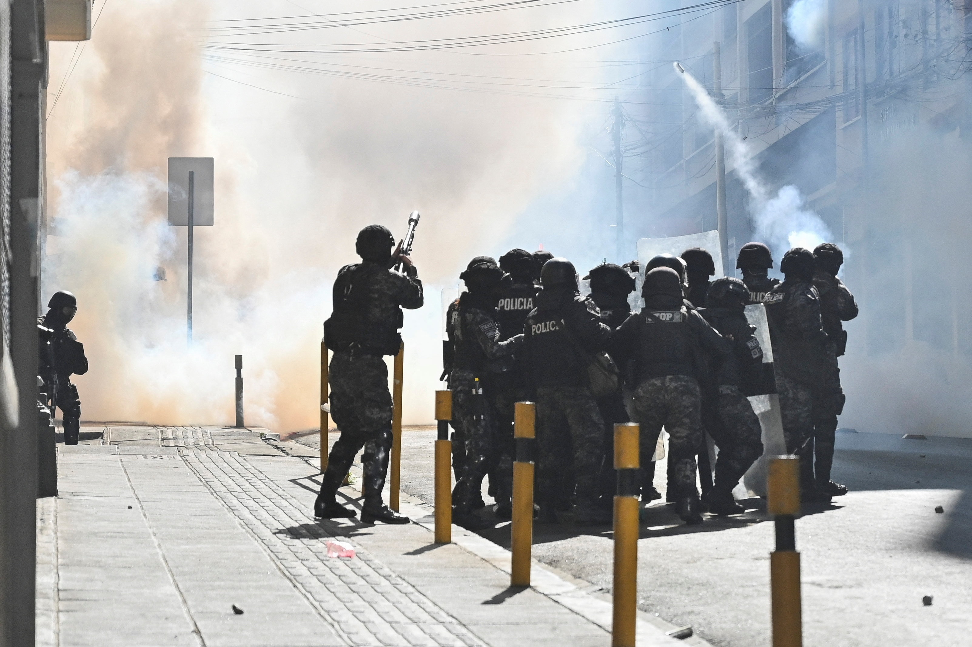 Police officers huddle together in a street and fire tear gas. Clouds of smoke fill the air.