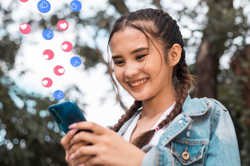 A smiling teenage Asian girl looks down at her phone as “heart” and “like” emojis float up from it.