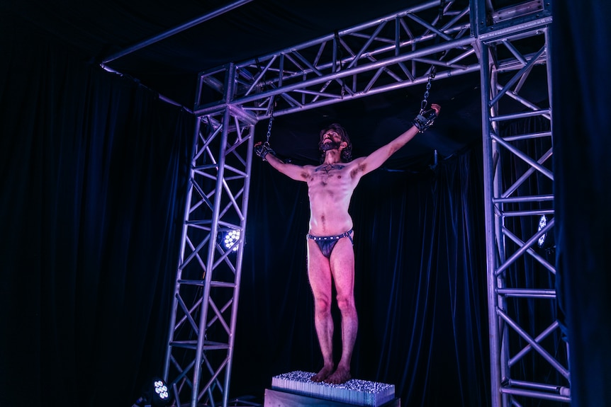 A middle-aged man in leather underwear stands upright on a raised platform with his hands chained to scaffolding above him