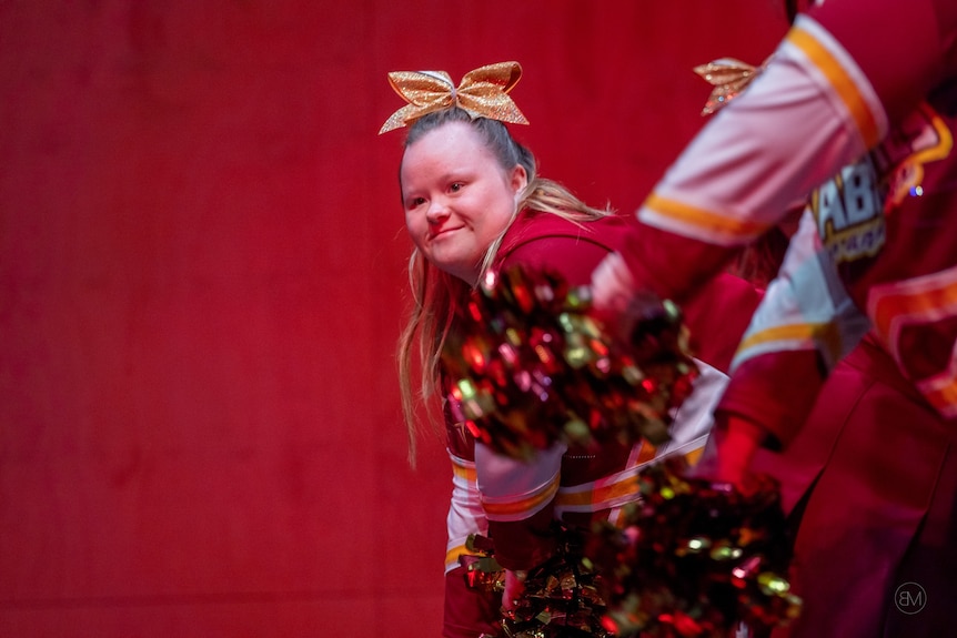 A dancer with a sparkly costume and yellow bow in her hair moves against a red backdrop
