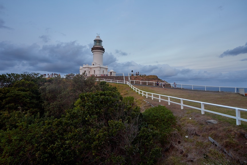 A lighthouse on a grassy hill stands tall amidst a cloudy sunset.