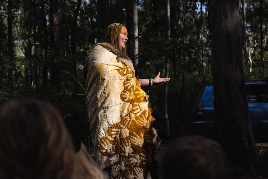 An Aboriginal woman, stands talking, in traditional clothing, with indigenous designs.