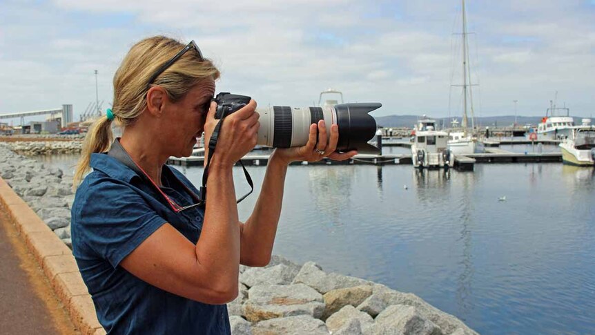 A woman with blonde hair and a blue shirt stands next to a marina and is photographed mid way through taking a photo herself.