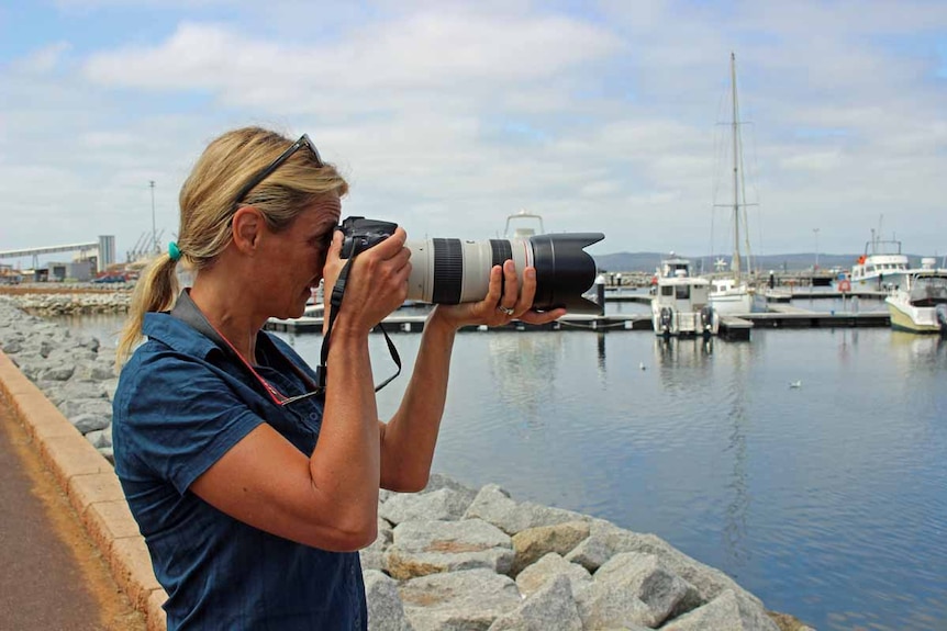 A woman with blonde hair and a blue shirt stands next to a marina and is photographed mid way through taking a photo herself.