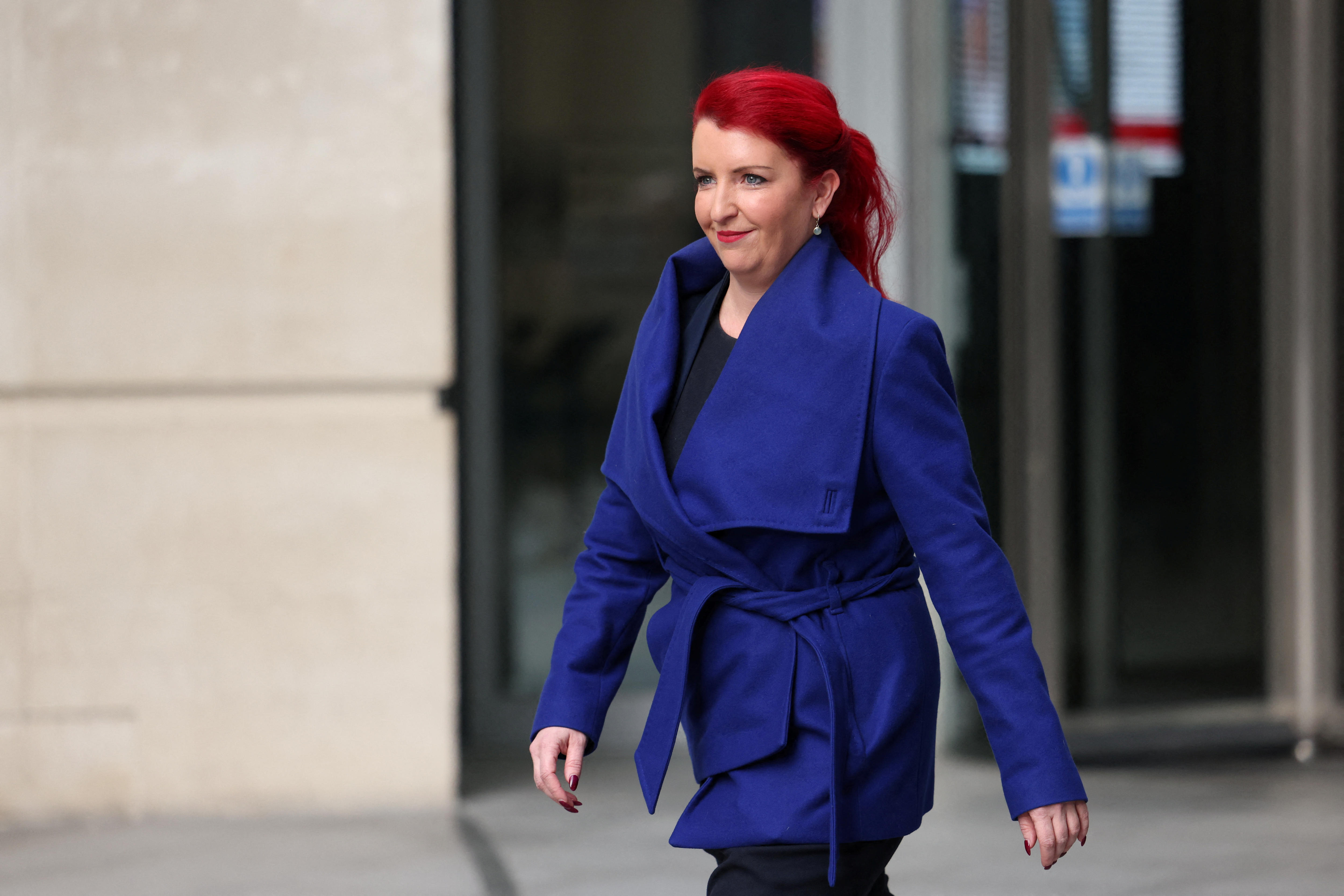 A young woman with bright red hair in a blue suit smiles as she walks past a sandstone building.