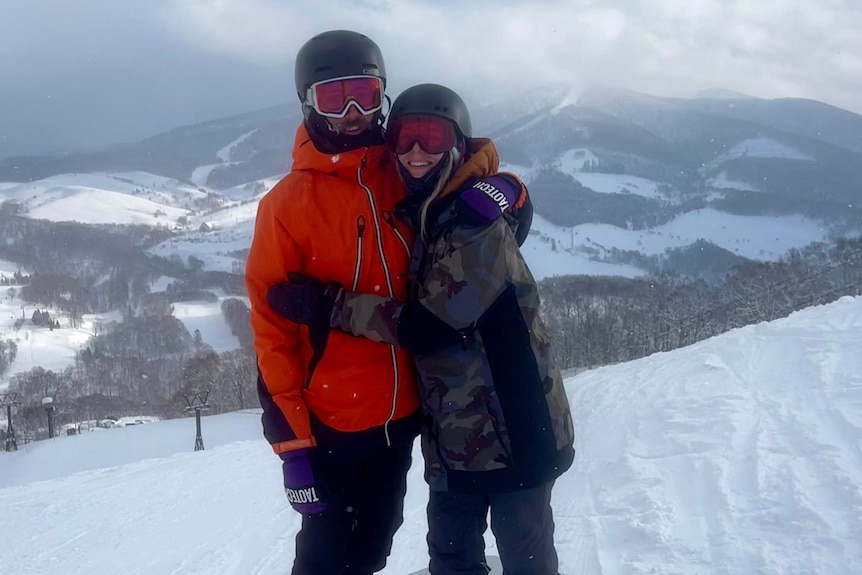 Couple in ski googles, and helmets, embrace in the snow smiling at the camera. Snowy mountains are visible behind them. 