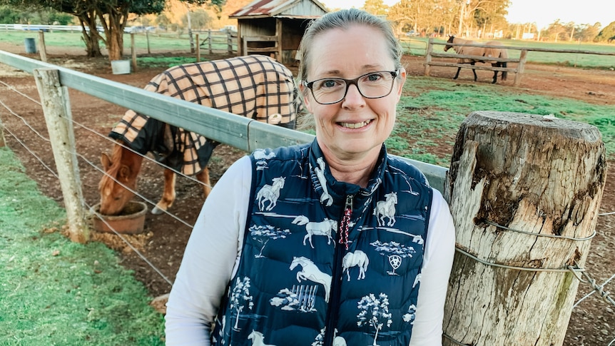 A woman with glasses standing in front a horse in a paddock