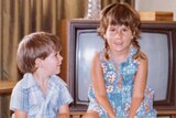 A young girl and boy sit on suitcases in a living room