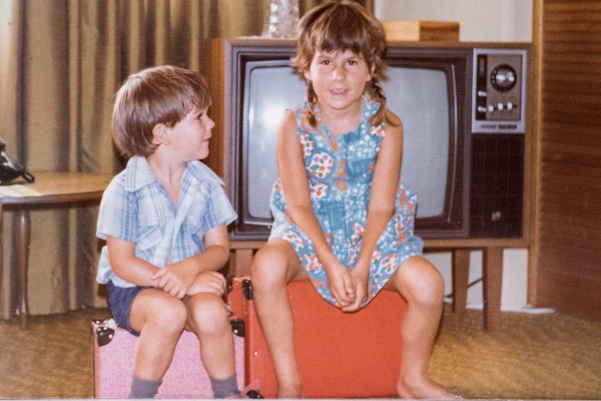 A young girl and boy sit on suitcases in a living room