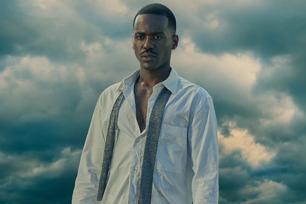 Actor Ncuti Gatwa in Doctor Who. He stands wearing a white shirt and undone tie. Behind him is a sky full of dark clouds.