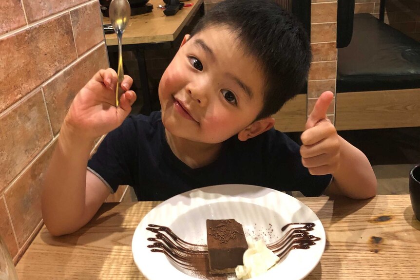 A young boy poses in a restaurant with a piece of chocolate cake.