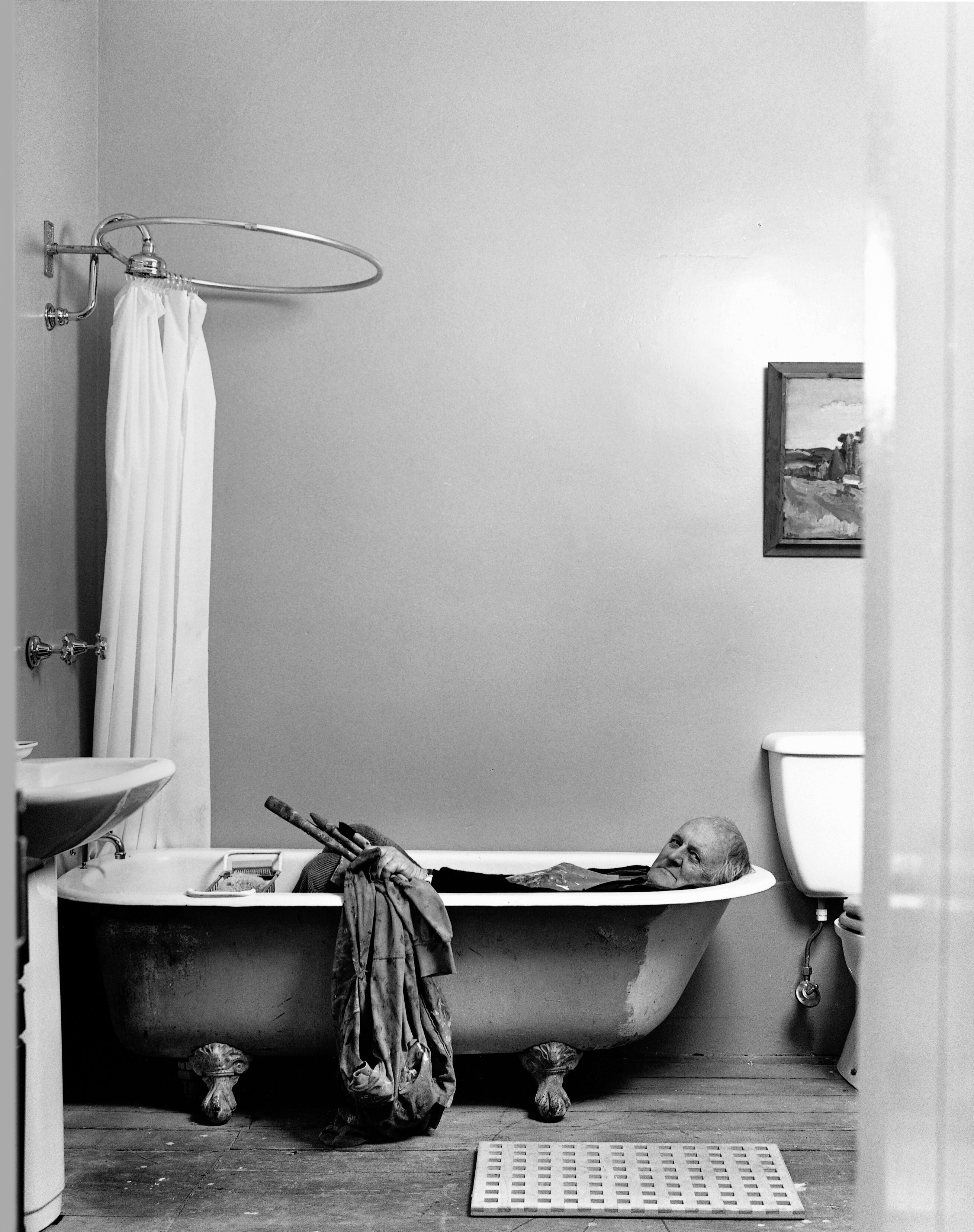 A black and white photo of a man in his early 70s lying in a claw foot bathtub holding pain brushes and canvas