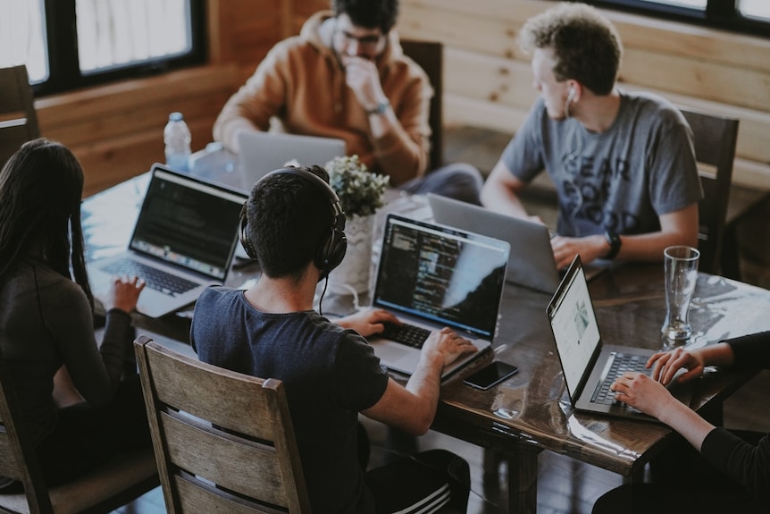 A group of young people sit at a table using laptops.