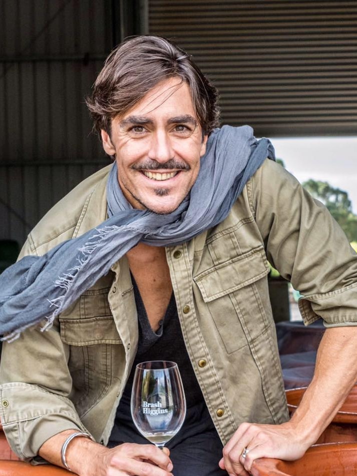 A dashing-looking, moustachioed man of indeterminate age, wearing a scarf and holding a wineglass.
