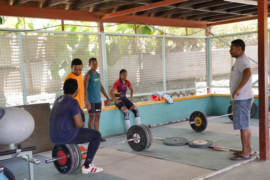 A woman sits down in front of weights as four men stand around her talking.