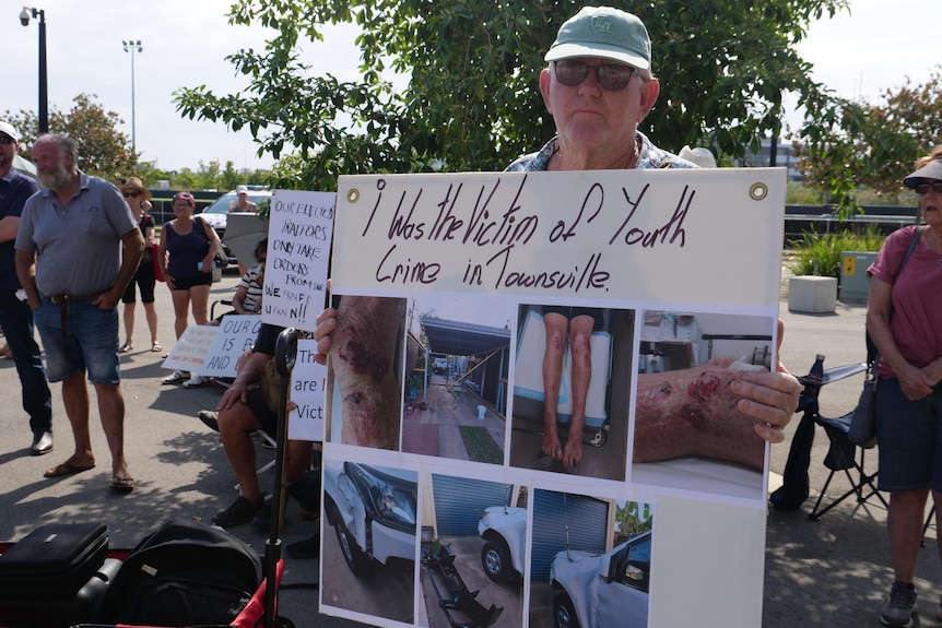 A man with a cap and sunglasses holds a sign showing pictures of his injuries. 