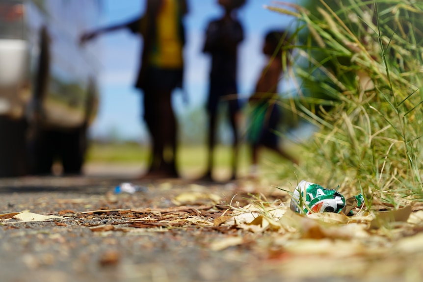 An empty can of VB lying on the ground on the side of a road, with the silhouettes of several people in the background.