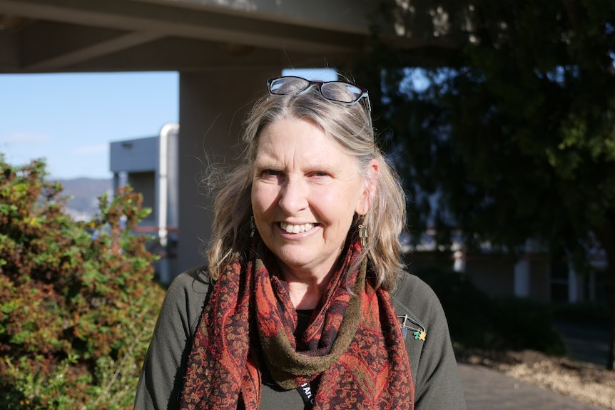 a woman wearing an orange scarf is standing outside, shrubs behind her, smiling at the camera