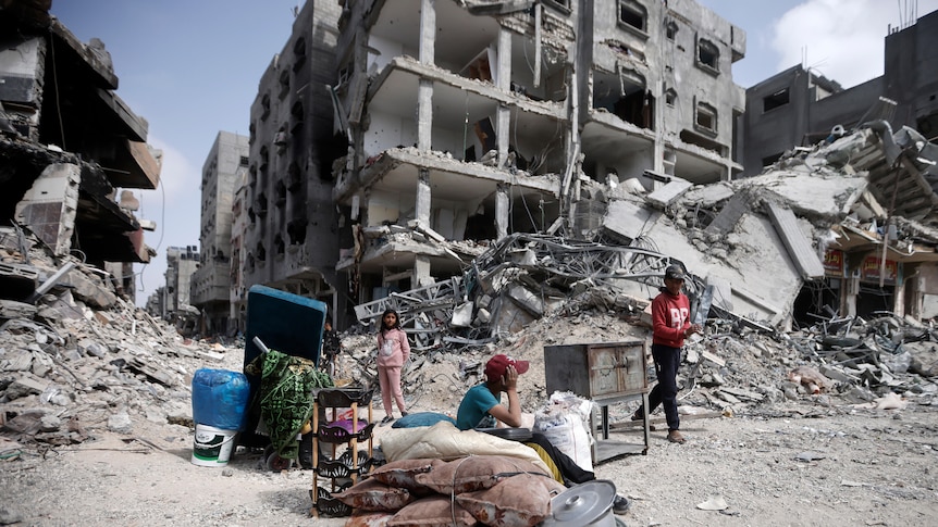 A wide shot of people sitting outsidte with some of their belongings, in front of destroyed buildings in the Gaza Strip