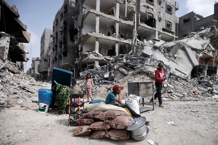 A wide shot of people sitting outsidte with some of their belongings, in front of destroyed buildings in the Gaza Strip