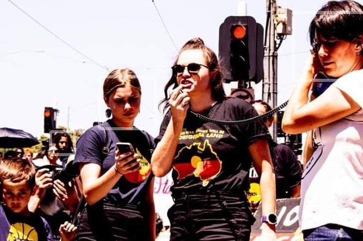 A woman wears an t-shirt with an indigenous flag and yells into a microphone at a protest