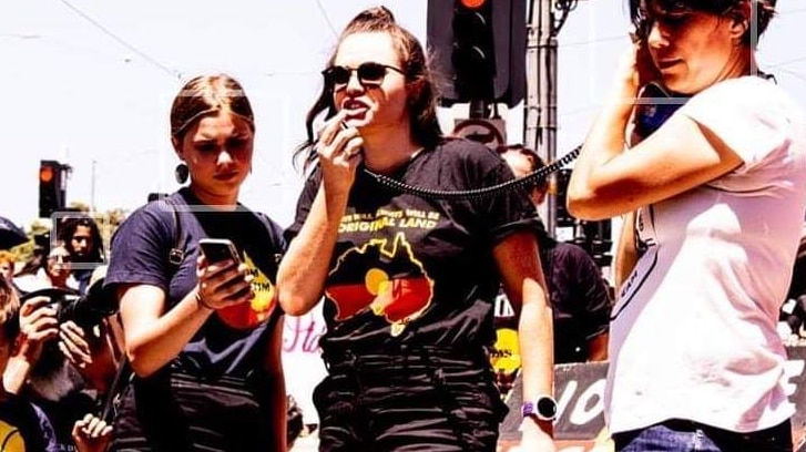 A woman wears an t-shirt with an indigenous flag and yells into a microphone at a protest