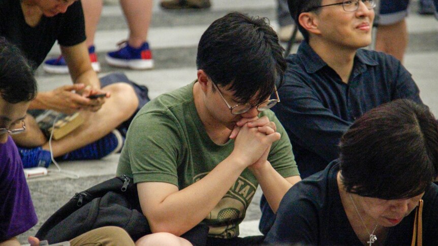 A man in a green t-shirt bows his head in prayer while sitting on the floor
