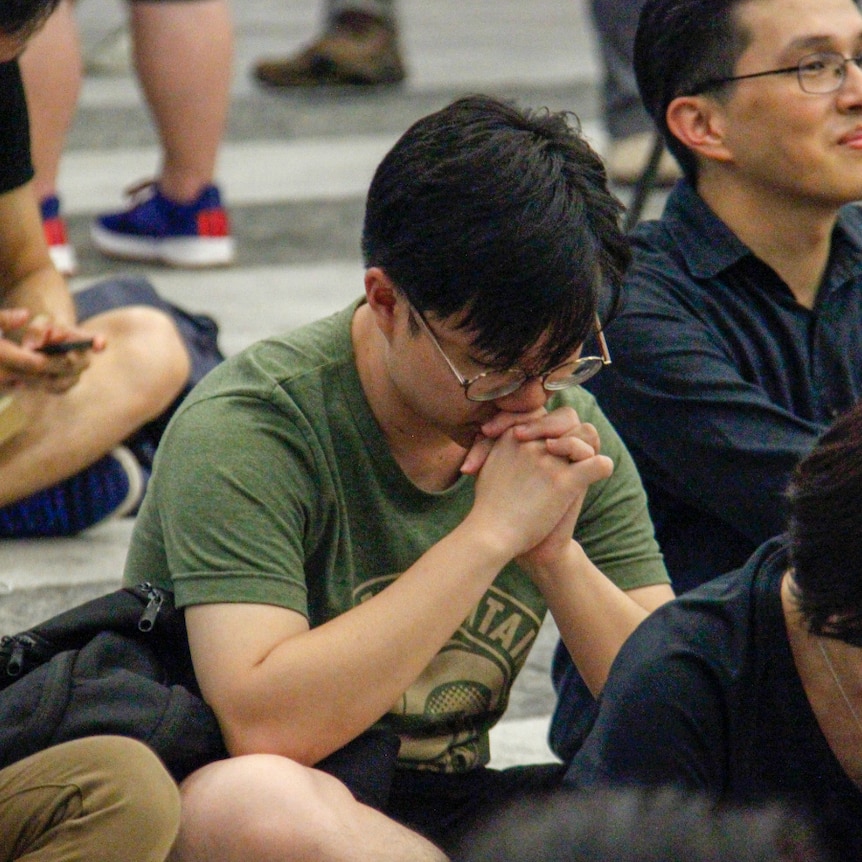 A man in a green t-shirt bows his head in prayer while sitting on the floor