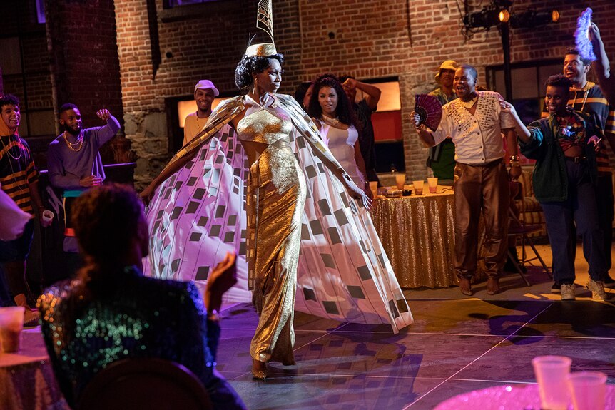 A beautiful Black drag queen showing off her resplendent gold outfit surrounded by cheering people in the TV series Pose