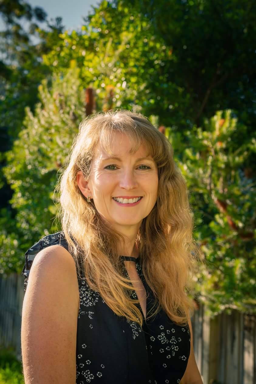 Dr Gribble stands facing the camera wearing a navy blue dress with trees in the background