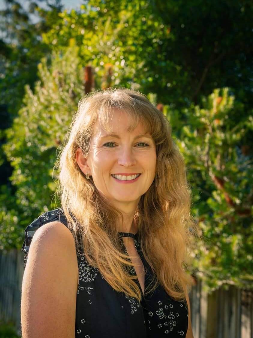 A woman with long blonde hair and blue eyes smiles at the camera. Behind her are green trees