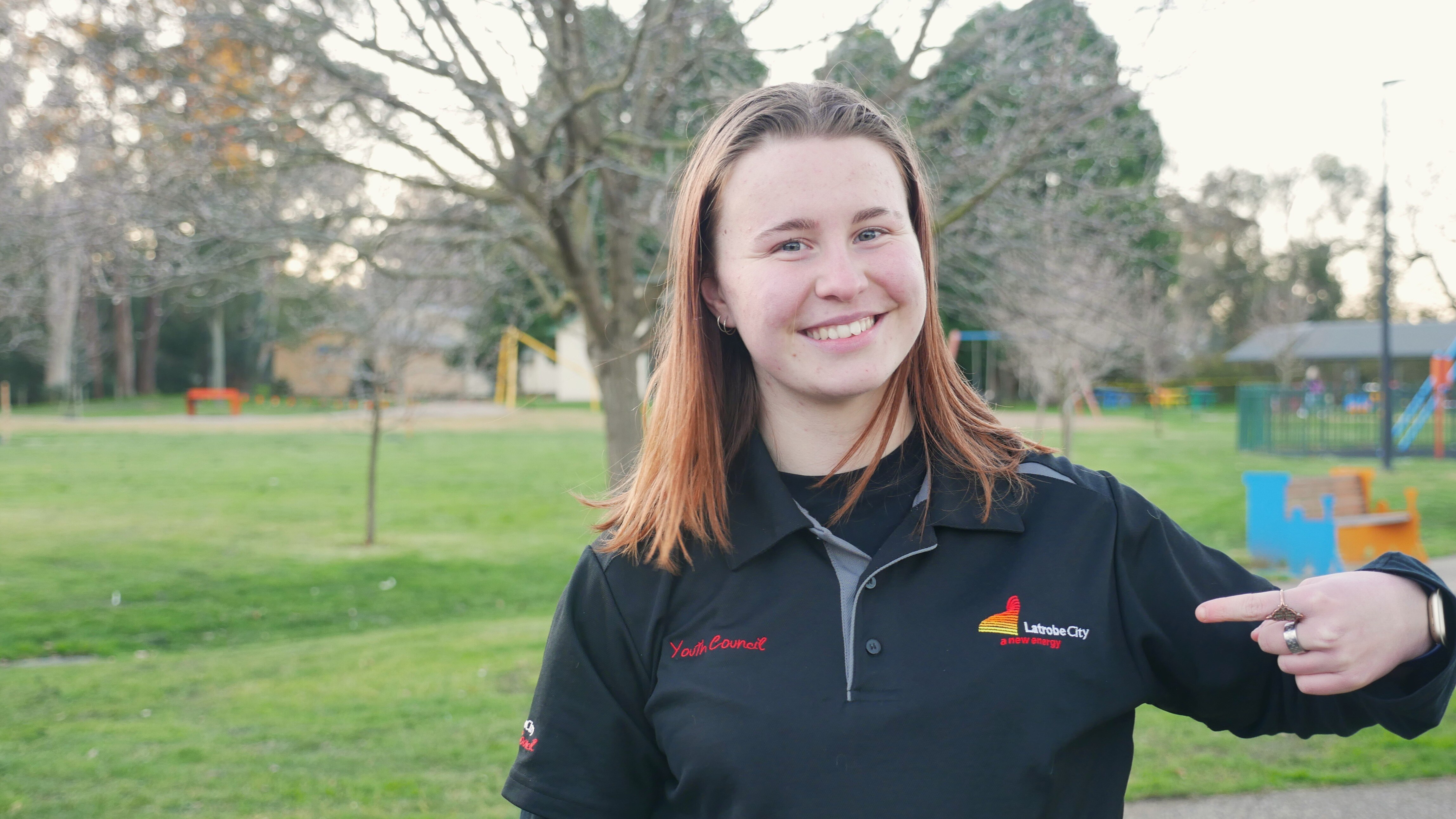 Young woman with shoulder length brown hair standing outside in a park, grass, bare trees, smiling pointing to her tshirt logo.